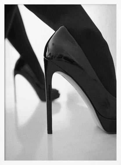 A close-up, black and white photograph of a person wearing shiny black patent leather stilettos and dark stockings. One shoe is in sharp focus in the foreground, highlighting its high arch and thin heel, while the other leg and shoe are softly blurred in the background on a reflective white surface. Decor