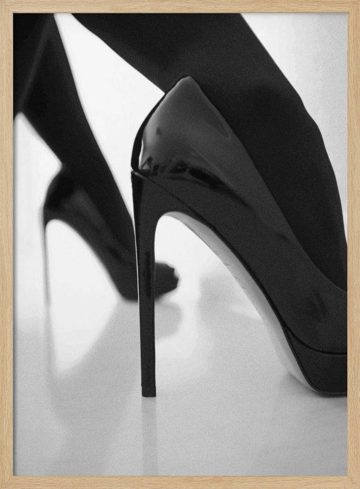 A close-up, black and white photograph of a person wearing shiny black patent leather stilettos and dark stockings. One shoe is in sharp focus in the foreground, highlighting its high arch and thin heel, while the other leg and shoe are softly blurred in the background on a reflective white surface. Decor