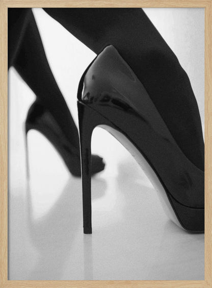 A close-up, black and white photograph of a person wearing shiny black patent leather stilettos and dark stockings. One shoe is in sharp focus in the foreground, highlighting its high arch and thin heel, while the other leg and shoe are softly blurred in the background on a reflective white surface. Decor