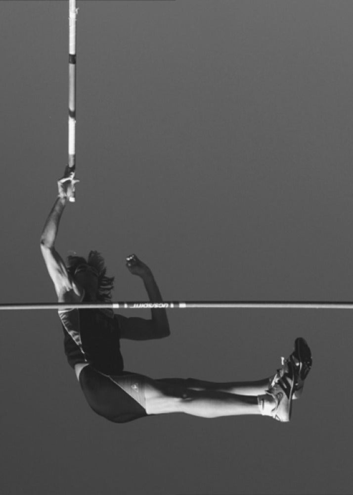 A dramatic black and white photograph of a male pole vaulter in mid-air, successfully clearing the horizontal bar. The athlete's body is parallel to the ground, with one arm still holding the vertical pole, against a solid gray sky. The image is enclosed in a simple black frame. Artwork