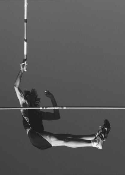 A dramatic black and white photograph of a male pole vaulter in mid-air, successfully clearing the horizontal bar. The athlete's body is parallel to the ground, with one arm still holding the vertical pole, against a solid gray sky. The image is enclosed in a simple black frame. Artwork