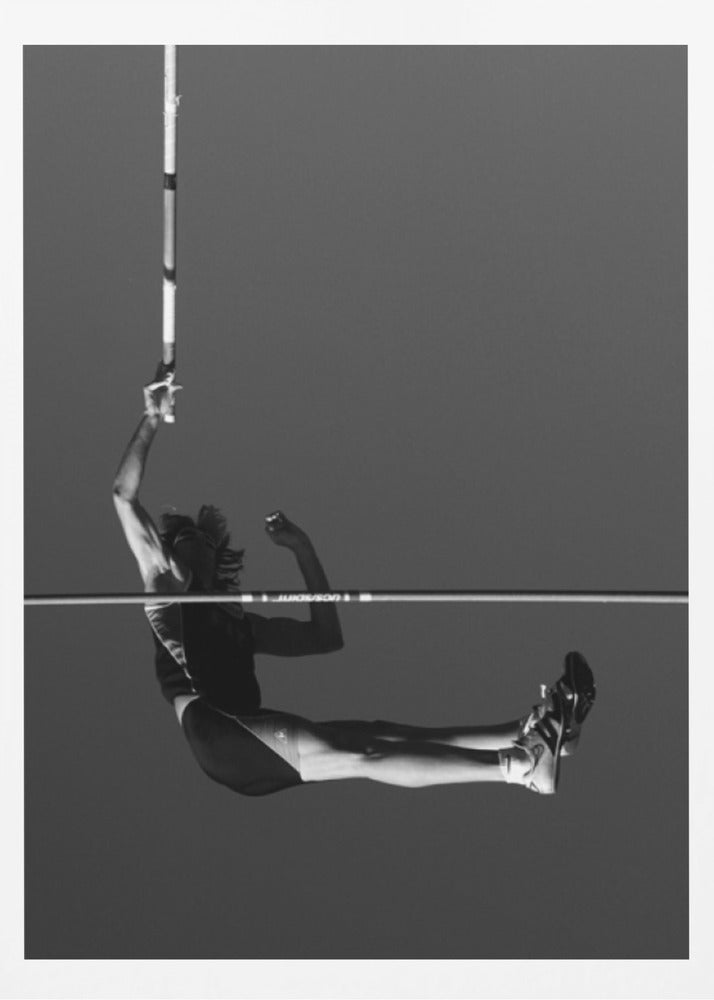 A dramatic black and white photograph of a male pole vaulter in mid-air, successfully clearing the horizontal bar. The athlete's body is parallel to the ground, with one arm still holding the vertical pole, against a solid gray sky. The image is enclosed in a simple black frame. Artwork