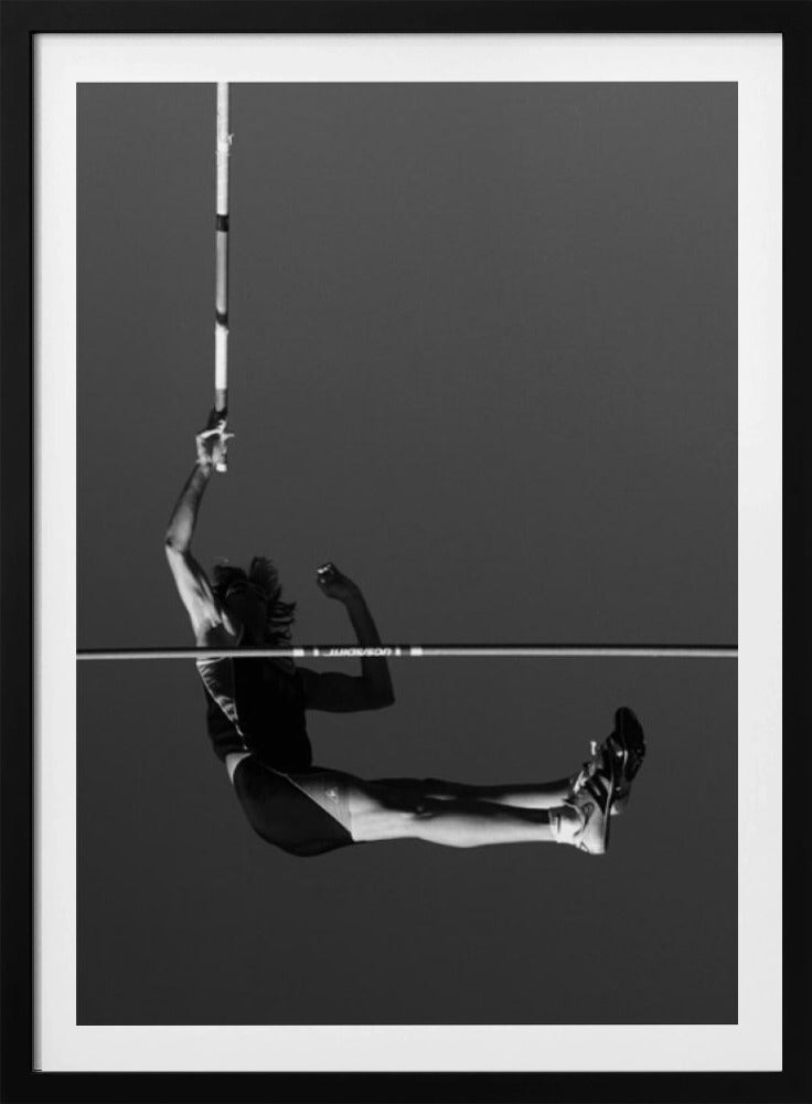 A dramatic black and white photograph of a male pole vaulter in mid-air, successfully clearing the horizontal bar. The athlete's body is parallel to the ground, with one arm still holding the vertical pole, against a solid gray sky. The image is enclosed in a simple black frame. Artwork