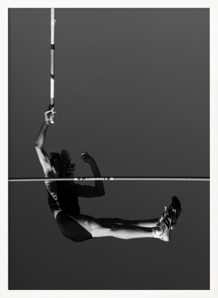 A dramatic black and white photograph of a male pole vaulter in mid-air, successfully clearing the horizontal bar. The athlete's body is parallel to the ground, with one arm still holding the vertical pole, against a solid gray sky. The image is enclosed in a simple black frame. Artwork