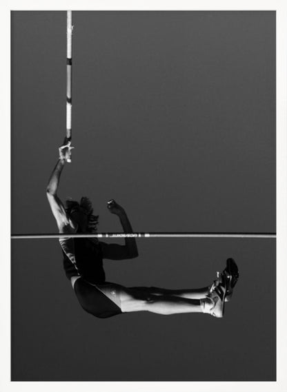 A dramatic black and white photograph of a male pole vaulter in mid-air, successfully clearing the horizontal bar. The athlete's body is parallel to the ground, with one arm still holding the vertical pole, against a solid gray sky. The image is enclosed in a simple black frame. Artwork
