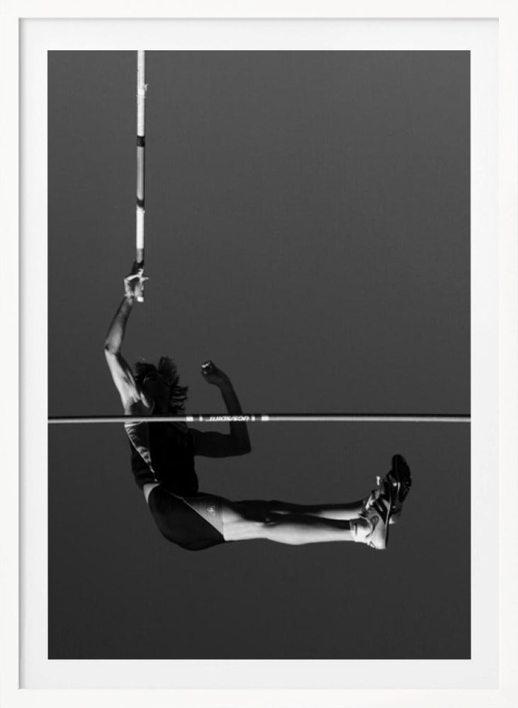 A dramatic black and white photograph of a male pole vaulter in mid-air, successfully clearing the horizontal bar. The athlete's body is parallel to the ground, with one arm still holding the vertical pole, against a solid gray sky. The image is enclosed in a simple black frame. Artwork