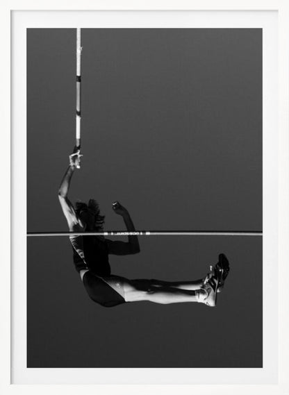 A dramatic black and white photograph of a male pole vaulter in mid-air, successfully clearing the horizontal bar. The athlete's body is parallel to the ground, with one arm still holding the vertical pole, against a solid gray sky. The image is enclosed in a simple black frame. Artwork