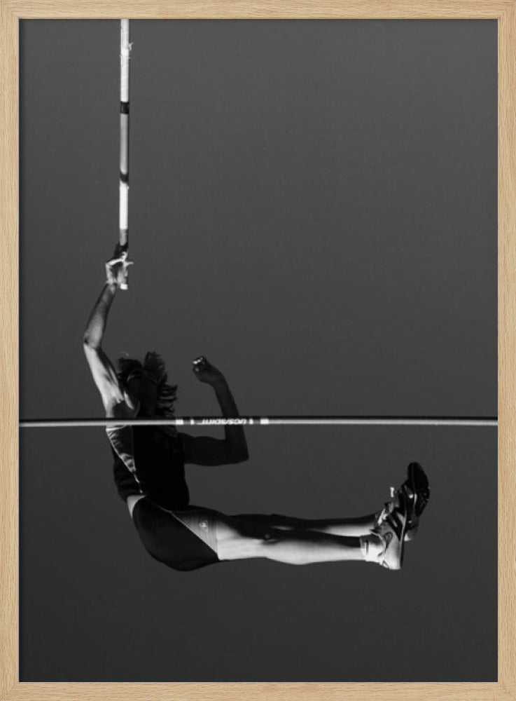 A dramatic black and white photograph of a male pole vaulter in mid-air, successfully clearing the horizontal bar. The athlete's body is parallel to the ground, with one arm still holding the vertical pole, against a solid gray sky. The image is enclosed in a simple black frame. Artwork