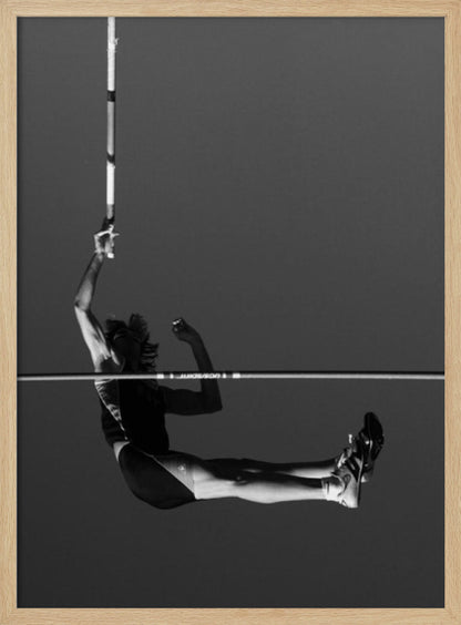 A dramatic black and white photograph of a male pole vaulter in mid-air, successfully clearing the horizontal bar. The athlete's body is parallel to the ground, with one arm still holding the vertical pole, against a solid gray sky. The image is enclosed in a simple black frame. Artwork