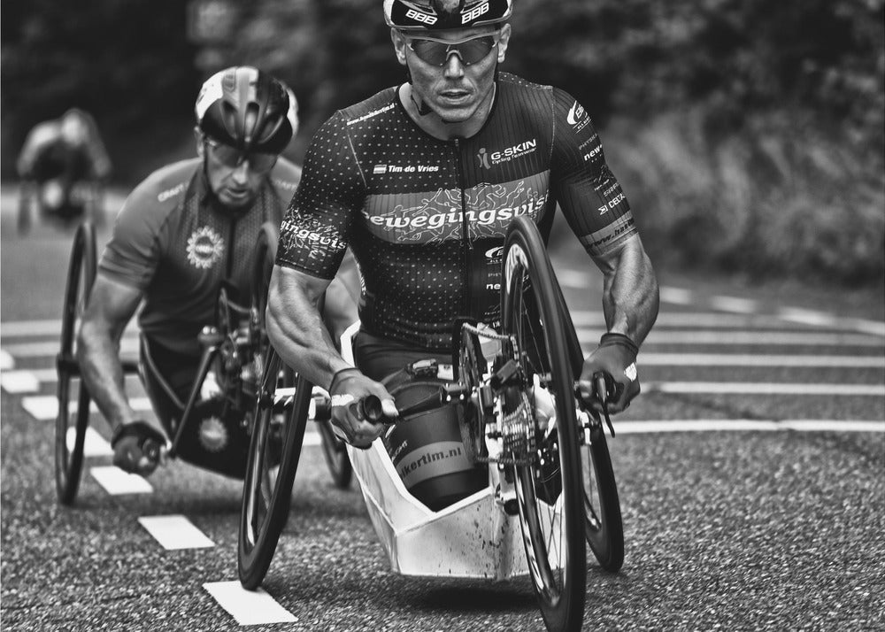 A powerful black and white photograph of two male athletes competing in a wheelchair race. The focused athlete in the foreground shows intense determination on his face, his muscular arms propelling the specialized racing wheelchair forward on an asphalt road. A second racer is visible in the background. Decor