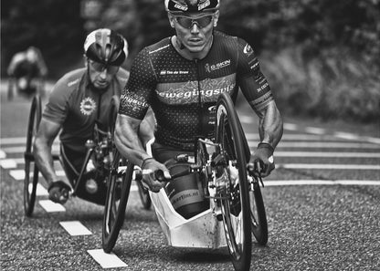 A powerful black and white photograph of two male athletes competing in a wheelchair race. The focused athlete in the foreground shows intense determination on his face, his muscular arms propelling the specialized racing wheelchair forward on an asphalt road. A second racer is visible in the background. Decor