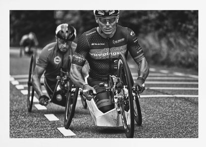 A powerful black and white photograph of two male athletes competing in a wheelchair race. The focused athlete in the foreground shows intense determination on his face, his muscular arms propelling the specialized racing wheelchair forward on an asphalt road. A second racer is visible in the background. Decor