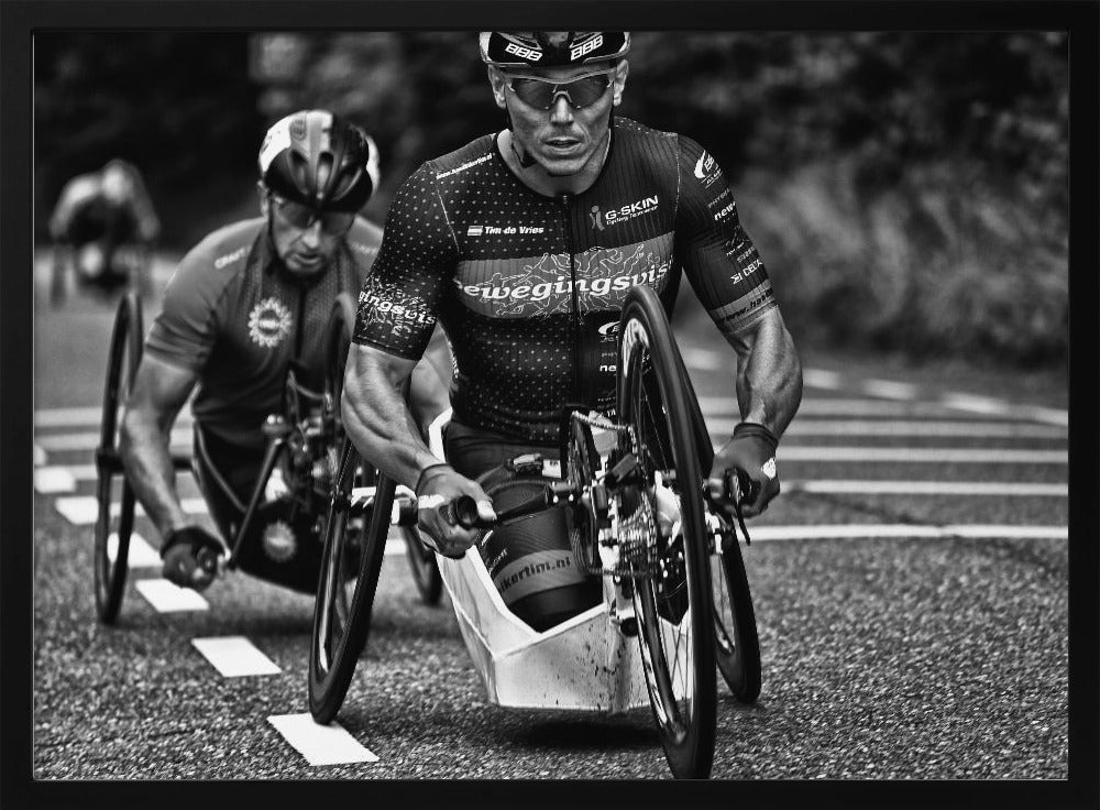 A powerful black and white photograph of two male athletes competing in a wheelchair race. The focused athlete in the foreground shows intense determination on his face, his muscular arms propelling the specialized racing wheelchair forward on an asphalt road. A second racer is visible in the background. Decor