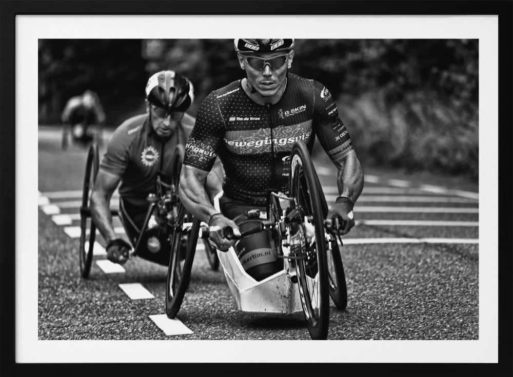 A powerful black and white photograph of two male athletes competing in a wheelchair race. The focused athlete in the foreground shows intense determination on his face, his muscular arms propelling the specialized racing wheelchair forward on an asphalt road. A second racer is visible in the background. Decor