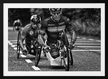 A powerful black and white photograph of two male athletes competing in a wheelchair race. The focused athlete in the foreground shows intense determination on his face, his muscular arms propelling the specialized racing wheelchair forward on an asphalt road. A second racer is visible in the background. Decor