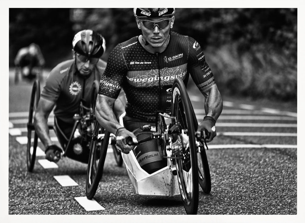 A powerful black and white photograph of two male athletes competing in a wheelchair race. The focused athlete in the foreground shows intense determination on his face, his muscular arms propelling the specialized racing wheelchair forward on an asphalt road. A second racer is visible in the background. Decor