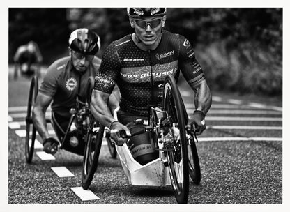 A powerful black and white photograph of two male athletes competing in a wheelchair race. The focused athlete in the foreground shows intense determination on his face, his muscular arms propelling the specialized racing wheelchair forward on an asphalt road. A second racer is visible in the background. Decor
