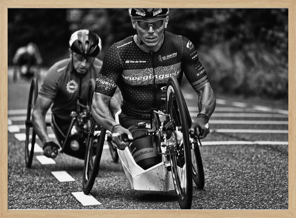 A powerful black and white photograph of two male athletes competing in a wheelchair race. The focused athlete in the foreground shows intense determination on his face, his muscular arms propelling the specialized racing wheelchair forward on an asphalt road. A second racer is visible in the background. Decor