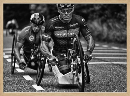 A powerful black and white photograph of two male athletes competing in a wheelchair race. The focused athlete in the foreground shows intense determination on his face, his muscular arms propelling the specialized racing wheelchair forward on an asphalt road. A second racer is visible in the background. Decor