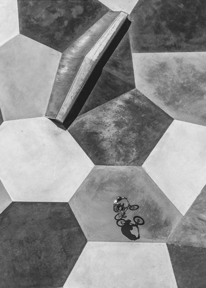 A high-angle, black and white photograph of a BMX rider in a skatepark with a geometric pattern. The ground consists of large, interlocking hexagons in various shades of grey, and the rider is captured mid-action on one of them, with a sharp shadow cast beside them. Artwork