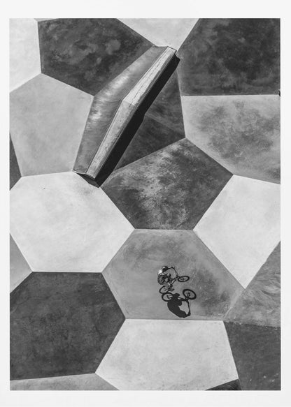 A high-angle, black and white photograph of a BMX rider in a skatepark with a geometric pattern. The ground consists of large, interlocking hexagons in various shades of grey, and the rider is captured mid-action on one of them, with a sharp shadow cast beside them. Artwork