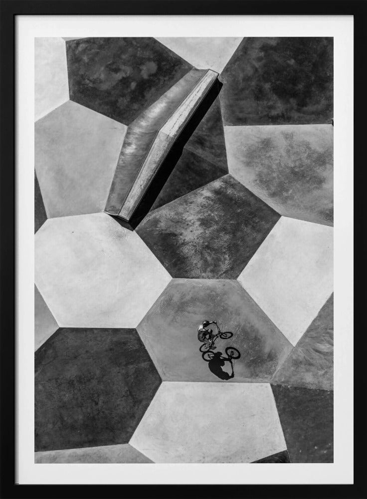 A high-angle, black and white photograph of a BMX rider in a skatepark with a geometric pattern. The ground consists of large, interlocking hexagons in various shades of grey, and the rider is captured mid-action on one of them, with a sharp shadow cast beside them. Artwork
