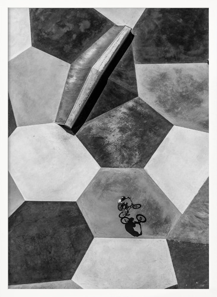 A high-angle, black and white photograph of a BMX rider in a skatepark with a geometric pattern. The ground consists of large, interlocking hexagons in various shades of grey, and the rider is captured mid-action on one of them, with a sharp shadow cast beside them. Artwork