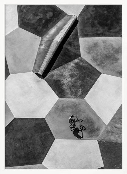 A high-angle, black and white photograph of a BMX rider in a skatepark with a geometric pattern. The ground consists of large, interlocking hexagons in various shades of grey, and the rider is captured mid-action on one of them, with a sharp shadow cast beside them. Artwork