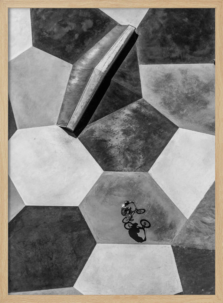 A high-angle, black and white photograph of a BMX rider in a skatepark with a geometric pattern. The ground consists of large, interlocking hexagons in various shades of grey, and the rider is captured mid-action on one of them, with a sharp shadow cast beside them. Artwork