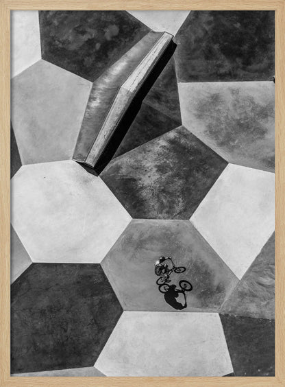 A high-angle, black and white photograph of a BMX rider in a skatepark with a geometric pattern. The ground consists of large, interlocking hexagons in various shades of grey, and the rider is captured mid-action on one of them, with a sharp shadow cast beside them. Artwork