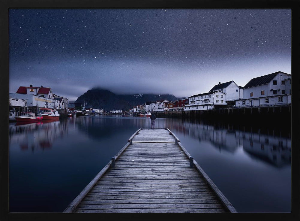 A long exposure photograph of a serene coastal town at night, viewed from a wooden pier that leads into calm, dark water. A lone person stands at the end of the pier, gazing at the town's reflected lights. Behind the village, a mountain looms under a breathtaking, star-filled sky. The image is presented within a silver-colored frame. Artwork