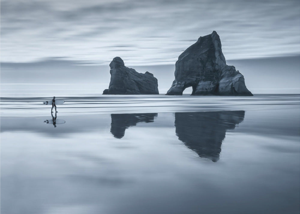A moody, blue-toned monochromatic photograph of a lone surfer walking on a wet, reflective beach. Two large rock formations stand in the calm ocean in the background, their reflections mirrored in the water. The sky is overcast, creating a serene and atmospheric scene, all enclosed in a silver frame. Decor