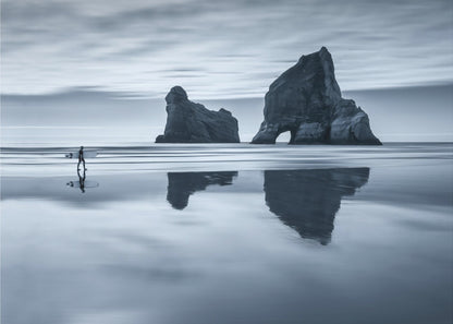 A moody, blue-toned monochromatic photograph of a lone surfer walking on a wet, reflective beach. Two large rock formations stand in the calm ocean in the background, their reflections mirrored in the water. The sky is overcast, creating a serene and atmospheric scene, all enclosed in a silver frame. Decor
