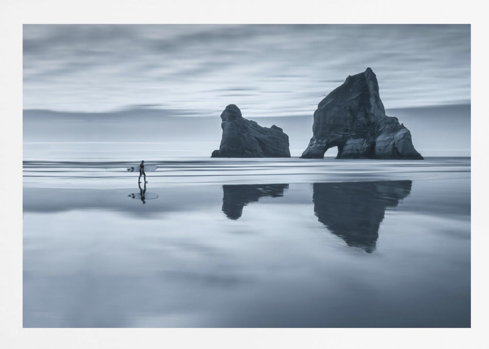 A moody, blue-toned monochromatic photograph of a lone surfer walking on a wet, reflective beach. Two large rock formations stand in the calm ocean in the background, their reflections mirrored in the water. The sky is overcast, creating a serene and atmospheric scene, all enclosed in a silver frame. Decor
