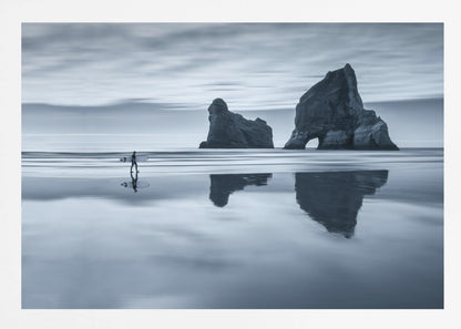 A moody, blue-toned monochromatic photograph of a lone surfer walking on a wet, reflective beach. Two large rock formations stand in the calm ocean in the background, their reflections mirrored in the water. The sky is overcast, creating a serene and atmospheric scene, all enclosed in a silver frame. Decor
