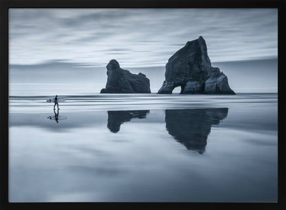 A moody, blue-toned monochromatic photograph of a lone surfer walking on a wet, reflective beach. Two large rock formations stand in the calm ocean in the background, their reflections mirrored in the water. The sky is overcast, creating a serene and atmospheric scene, all enclosed in a silver frame. Decor