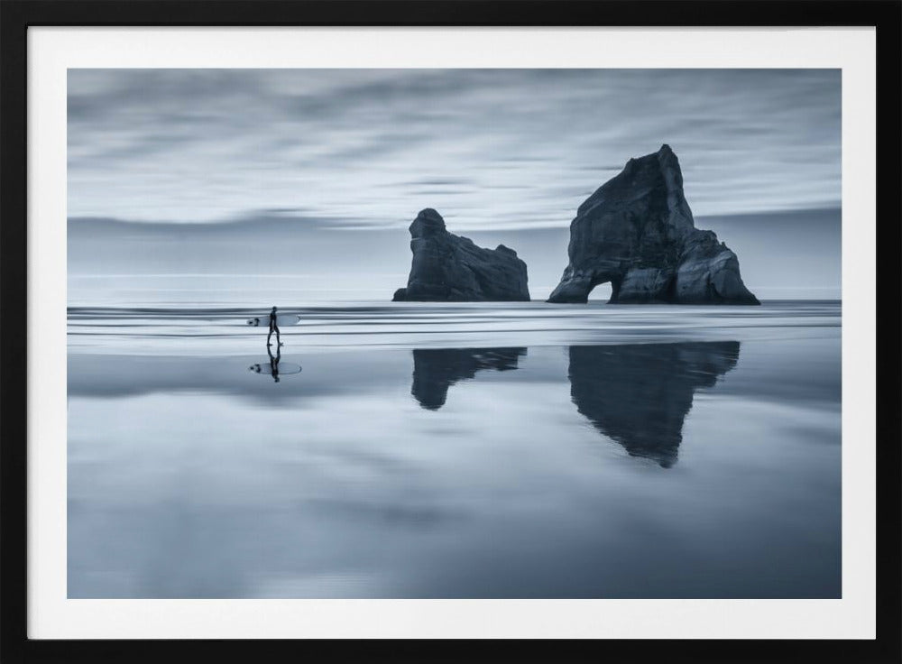 A moody, blue-toned monochromatic photograph of a lone surfer walking on a wet, reflective beach. Two large rock formations stand in the calm ocean in the background, their reflections mirrored in the water. The sky is overcast, creating a serene and atmospheric scene, all enclosed in a silver frame. Decor