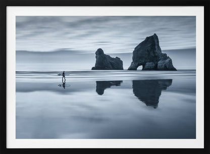 A moody, blue-toned monochromatic photograph of a lone surfer walking on a wet, reflective beach. Two large rock formations stand in the calm ocean in the background, their reflections mirrored in the water. The sky is overcast, creating a serene and atmospheric scene, all enclosed in a silver frame. Decor