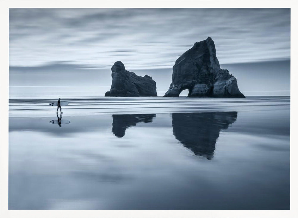 A moody, blue-toned monochromatic photograph of a lone surfer walking on a wet, reflective beach. Two large rock formations stand in the calm ocean in the background, their reflections mirrored in the water. The sky is overcast, creating a serene and atmospheric scene, all enclosed in a silver frame. Decor