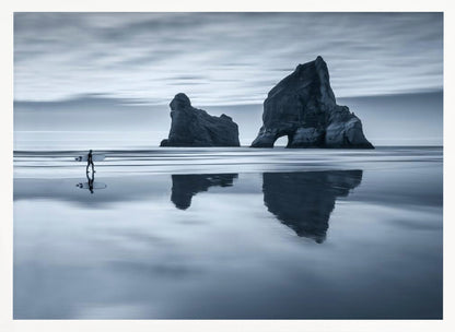 A moody, blue-toned monochromatic photograph of a lone surfer walking on a wet, reflective beach. Two large rock formations stand in the calm ocean in the background, their reflections mirrored in the water. The sky is overcast, creating a serene and atmospheric scene, all enclosed in a silver frame. Decor