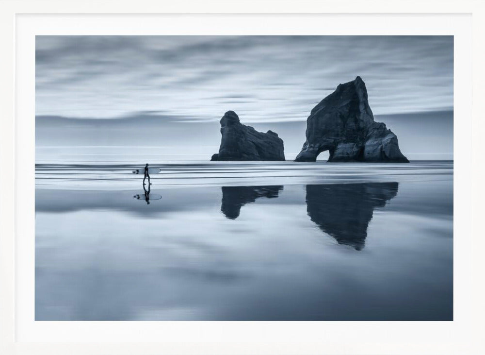 A moody, blue-toned monochromatic photograph of a lone surfer walking on a wet, reflective beach. Two large rock formations stand in the calm ocean in the background, their reflections mirrored in the water. The sky is overcast, creating a serene and atmospheric scene, all enclosed in a silver frame. Decor