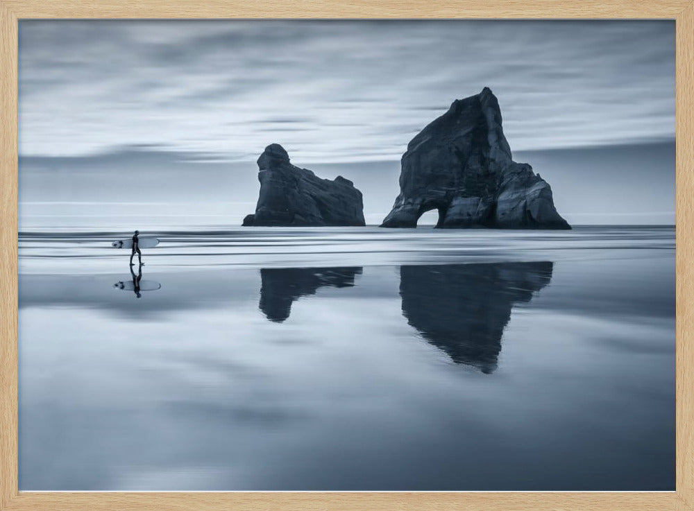A moody, blue-toned monochromatic photograph of a lone surfer walking on a wet, reflective beach. Two large rock formations stand in the calm ocean in the background, their reflections mirrored in the water. The sky is overcast, creating a serene and atmospheric scene, all enclosed in a silver frame. Decor