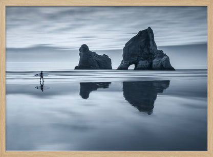 A moody, blue-toned monochromatic photograph of a lone surfer walking on a wet, reflective beach. Two large rock formations stand in the calm ocean in the background, their reflections mirrored in the water. The sky is overcast, creating a serene and atmospheric scene, all enclosed in a silver frame. Decor