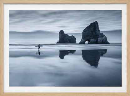 A moody, blue-toned monochromatic photograph of a lone surfer walking on a wet, reflective beach. Two large rock formations stand in the calm ocean in the background, their reflections mirrored in the water. The sky is overcast, creating a serene and atmospheric scene, all enclosed in a silver frame. Decor