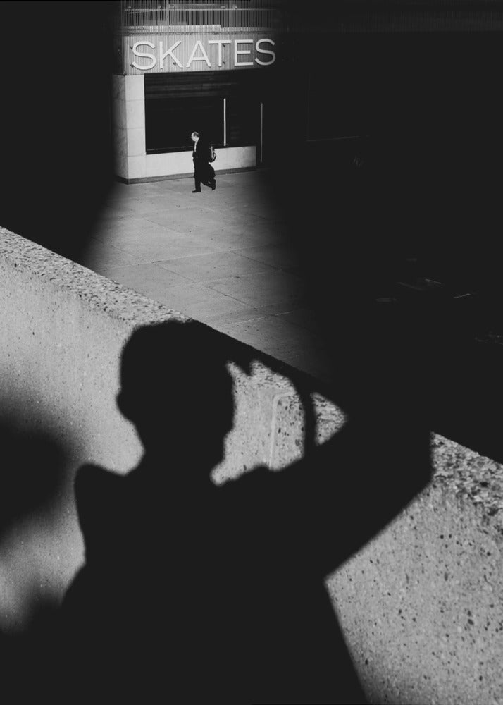 A high-contrast, black and white street photograph where the large shadow of the photographer is cast on a concrete barrier in the foreground. In the background, a lone person walks through a spotlit area of sidewalk in front of a building with an illuminated sign that says 'SKATES'. Poster