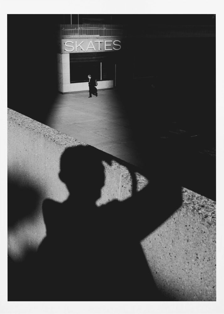 A high-contrast, black and white street photograph where the large shadow of the photographer is cast on a concrete barrier in the foreground. In the background, a lone person walks through a spotlit area of sidewalk in front of a building with an illuminated sign that says 'SKATES'. Poster