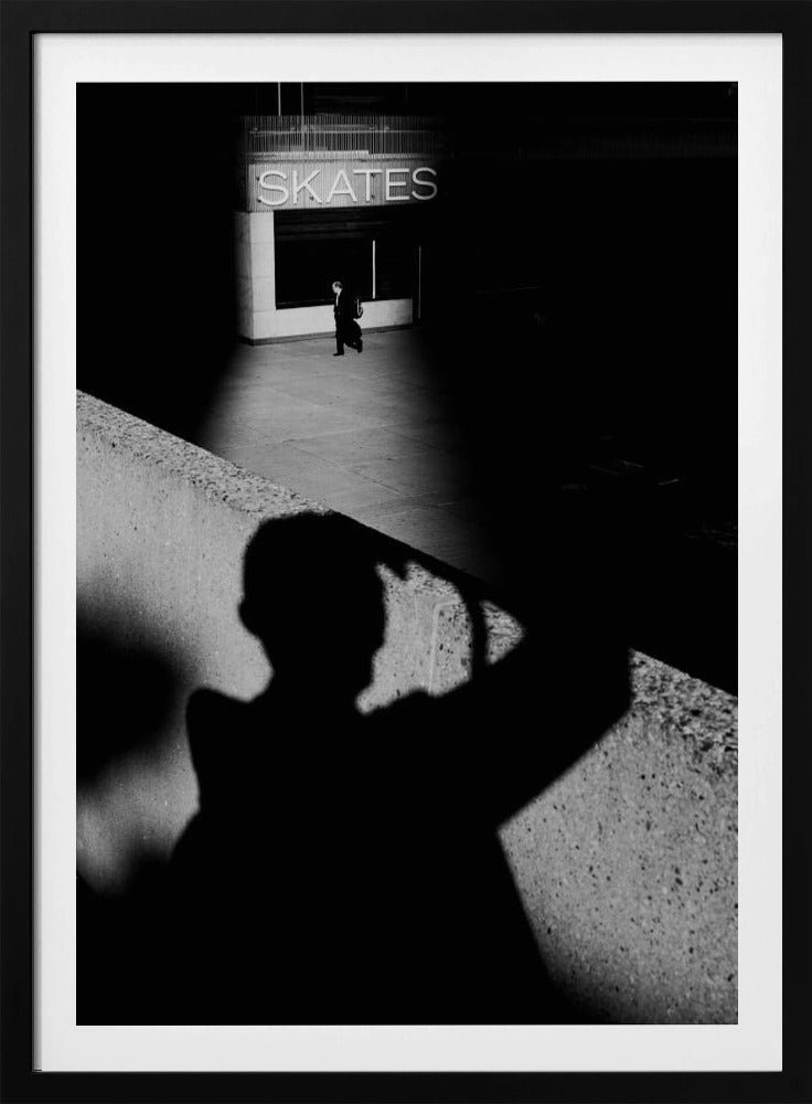 A high-contrast, black and white street photograph where the large shadow of the photographer is cast on a concrete barrier in the foreground. In the background, a lone person walks through a spotlit area of sidewalk in front of a building with an illuminated sign that says 'SKATES'. Poster