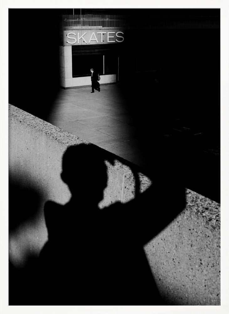 A high-contrast, black and white street photograph where the large shadow of the photographer is cast on a concrete barrier in the foreground. In the background, a lone person walks through a spotlit area of sidewalk in front of a building with an illuminated sign that says 'SKATES'. Poster