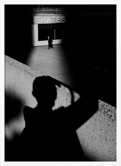 A high-contrast, black and white street photograph where the large shadow of the photographer is cast on a concrete barrier in the foreground. In the background, a lone person walks through a spotlit area of sidewalk in front of a building with an illuminated sign that says 'SKATES'. Poster