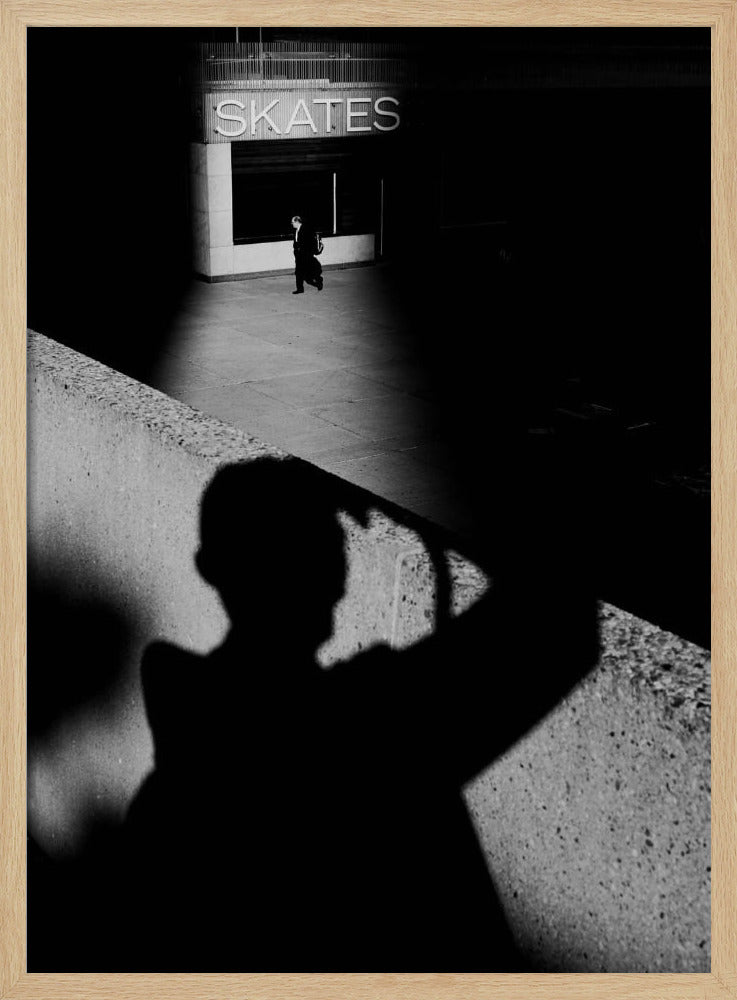 A high-contrast, black and white street photograph where the large shadow of the photographer is cast on a concrete barrier in the foreground. In the background, a lone person walks through a spotlit area of sidewalk in front of a building with an illuminated sign that says 'SKATES'. Poster